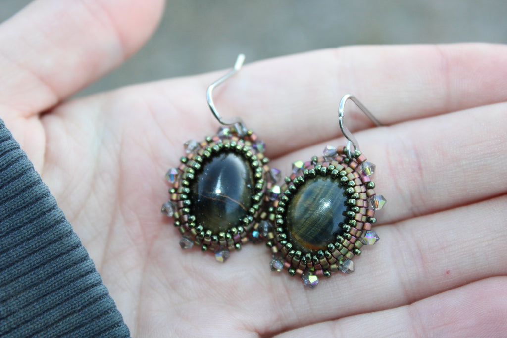 A pair of handmade beaded dangle earrings with oval tiger's eye stones and silver fishhook wires, held in a person's hand.